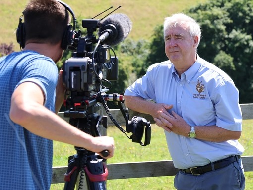 A man holding a camera filming an older man leaning against a fence